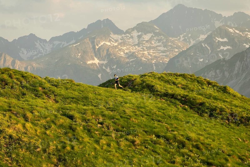 A lone runner is captured mid-stride on a lush green trail with majestic mountains in the background. The scene highlights the thrill and challenge of trail running in nature