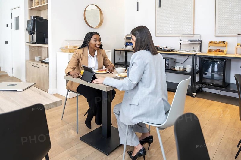 Two businesswomen are having breakfast and using a tablet in a hotel lobby during a business trip