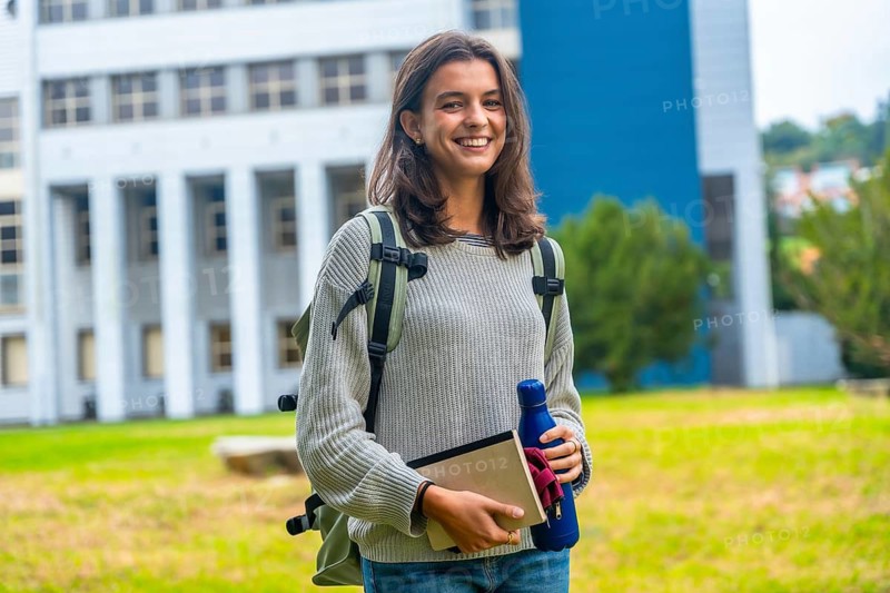Young female student smiling and looking at the camera, holding a notebook and water bottle while standing outdoors on a university campus with a building in the background