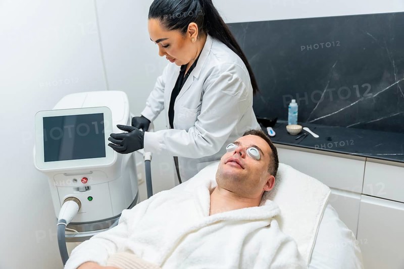 Man reclined on a white clinic bed wearing protective goggles while a beautician uses a modern laser device for facial skin rejuvenation and professional dermatology care