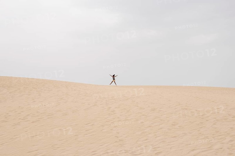 A lone individual joyfully jumps on a vast sand dune under a cloudy summer sky, capturing the essence of freedom and adventure in a serene desert landscape