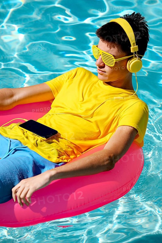A person in a yellow shirt, sunglasses, and headphones lounges on a pink inflatable float in a swimming pool. The scene evokes a carefree summer day and relaxation