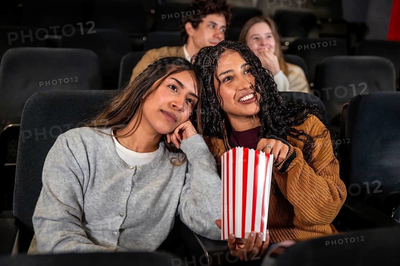 Two friends enjoying a movie at the cinema, one is eating popcorn while the other looks bored