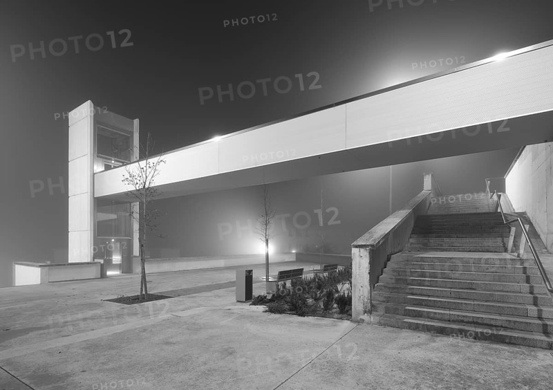 A modern urban elevator and connecting staircase are shrouded in dense mist under a glowing light, casting an eerie yet serene atmosphere