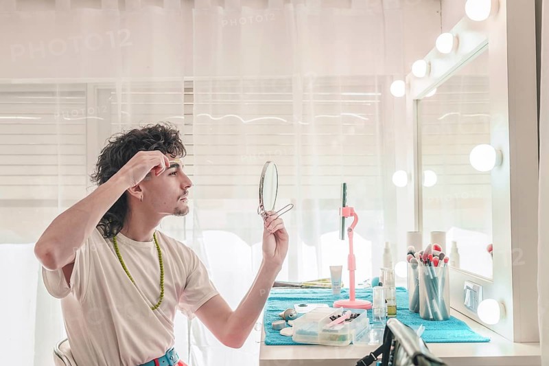 Young man plucking his eyebrows in a bedroom with a vanity mirror surrounded by led lights, showcasing a modern approach to beauty and self-care