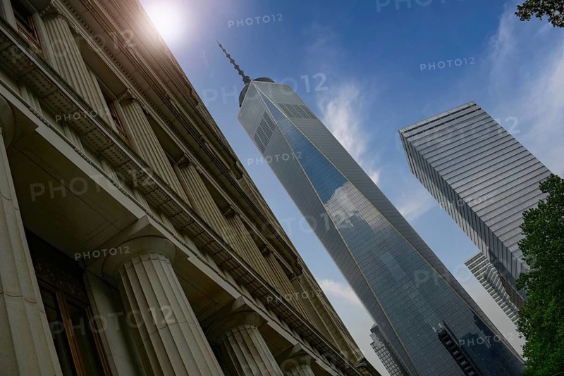 From below a beautiful perspective of Manhattan's iconic One World Trade Center against a clear blue sky, accompanied by neighboring modern buildings and historical architecture