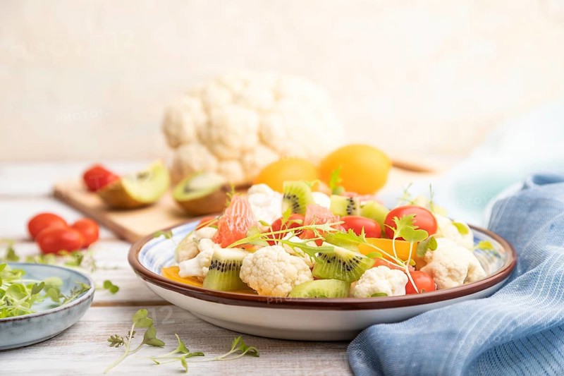 Vegetarian salad of cauliflower cabbage, kiwi, tomatoes, microgreen sprouts on white wooden background and blue linen textile. Side view, close up, selective focus