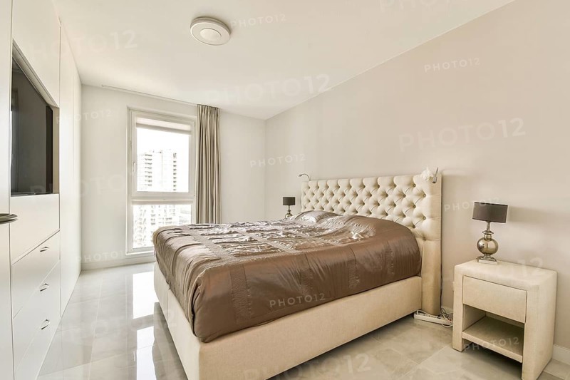 Modern minimalist bedroom showcasing a plush bed, elegant bedside lamps, and soft beige tones. Natural light spills through a large window, creating a serene atmosphere
