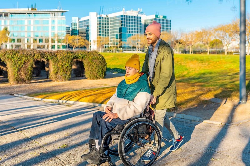 Young man pushing his disabled friend in a wheelchair while enjoying a sunny day together in a modern urban park