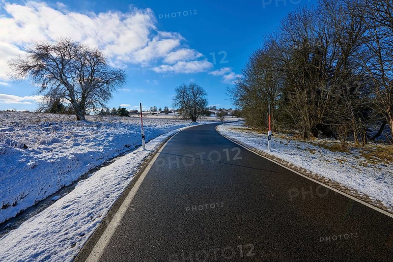 A sunny country road winds through a snow-covered landscape with bare trees and blue sky, winter, Oberelsbach, Lange Rhön, Rhön, Bavaria, Germany
