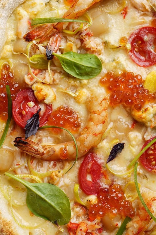 Close up view of a delectable gourmet pizza topped with succulent shrimp, vibrant red caviar, creamy mozzarella, sliced tomatoes, basil spinach, and savory crab meat, served on a crisp crust