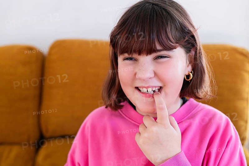 A cheerful young girl in a pink sweatshirt smiles and points to her missing milk tooth, seated in a cozy living room background