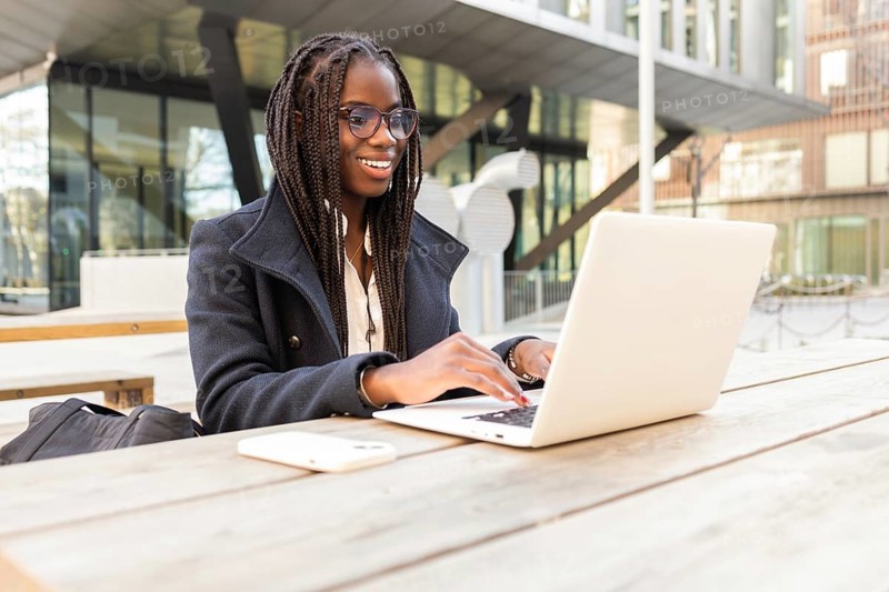 A young African American businesswoman with braids works on a laptop at an outdoor table in a casual setting