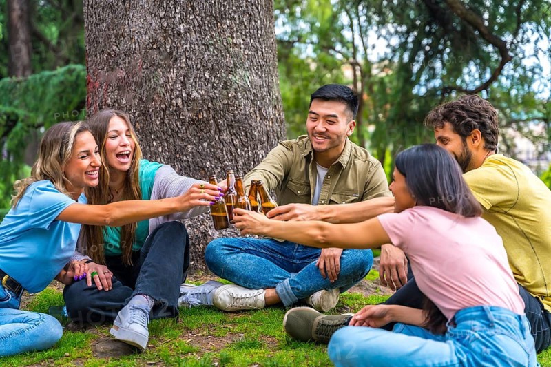 Group of multi-ethnic friends sitting in the city park talking next to a tree toasting with bottles of beers