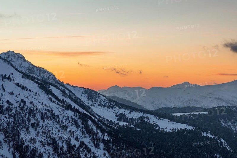 A serene snowy mountain landscape bathed in the warm glow of a sunset, showcasing a gradient of orange to blue skies above silhouetted peaks