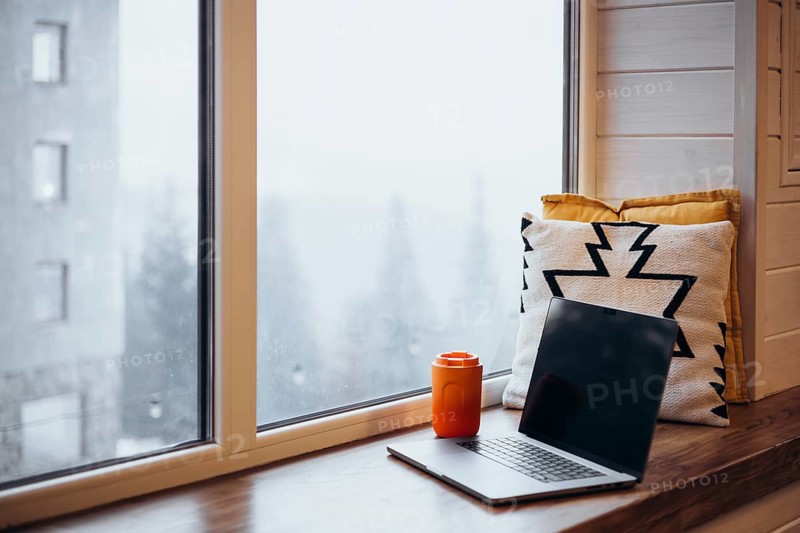 A peaceful remote work environment featuring a laptop and an orange thermos cup beside a snow-covered window, complemented by a cushion with bold patterns, creating a calming workspace at home
