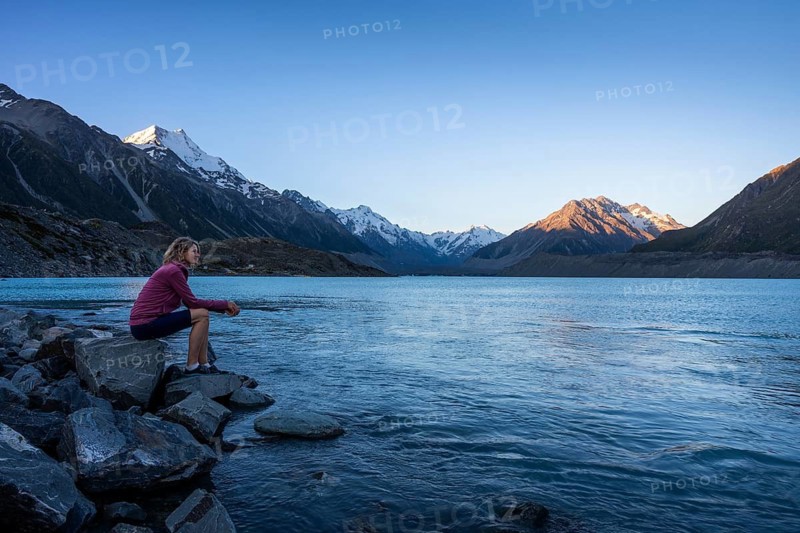 A woman is sitting on a rock at Tasman Lake. On the left, Mount Cook. Golden hour. Aoraki Mount Cook National Park, Canterbury, South Island, New Zealand