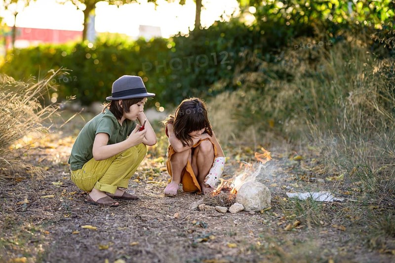 Two children, one in a hat, sit by a small campfire in a natural setting. Surrounded by trees and grass, they carefully observe the flames, experiencing outdoor adventure