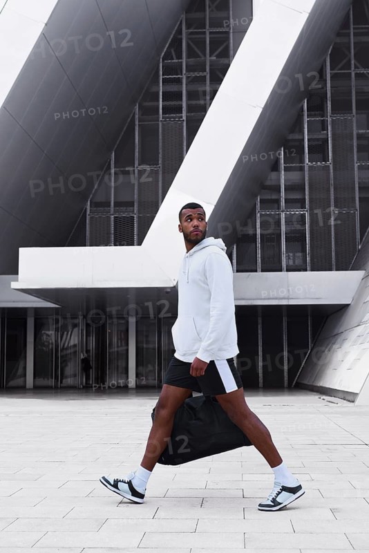 A confident black male athlete is captured in motion, wearing a hoodie and shorts in an urban setting. The modern architecture complements his dynamic presence