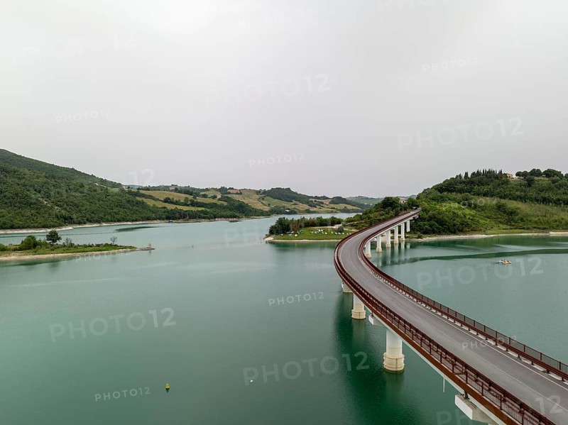 Aerial view of the scenic curved bridge spanning Castreccioni Lake in Italy. Calm turquoise waters and surrounding rolling hills create a peaceful natural and architectural landscape