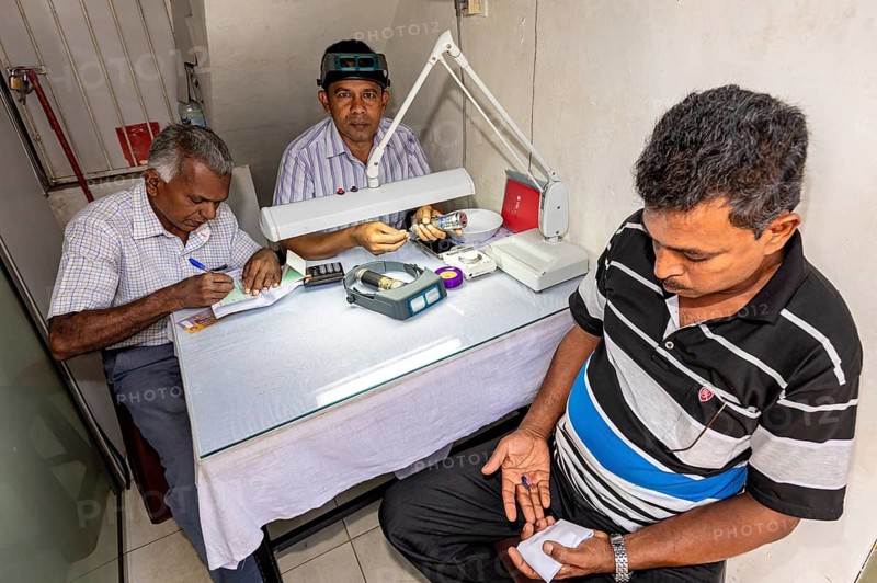 Three men work at a table, one writing while the other examines gemstones under a magnifying glass, The gemstone cutters in the city of Ratnapura in Sri Lanka