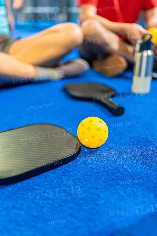 Pickleball paddles and a yellow wiffle ball resting on a blue court surface, with players relaxing in the background, depicting a moment of pause during active sports