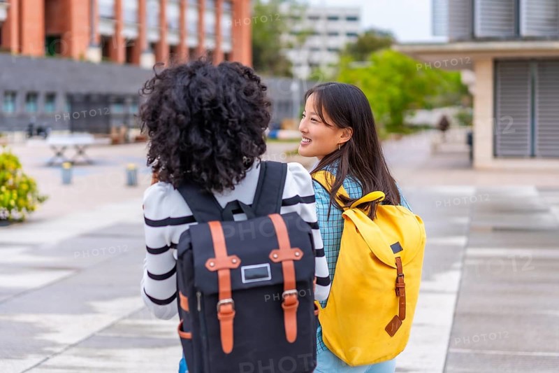 Rear view of two multi-ethnic female students walking along an urban campus talking and smiling