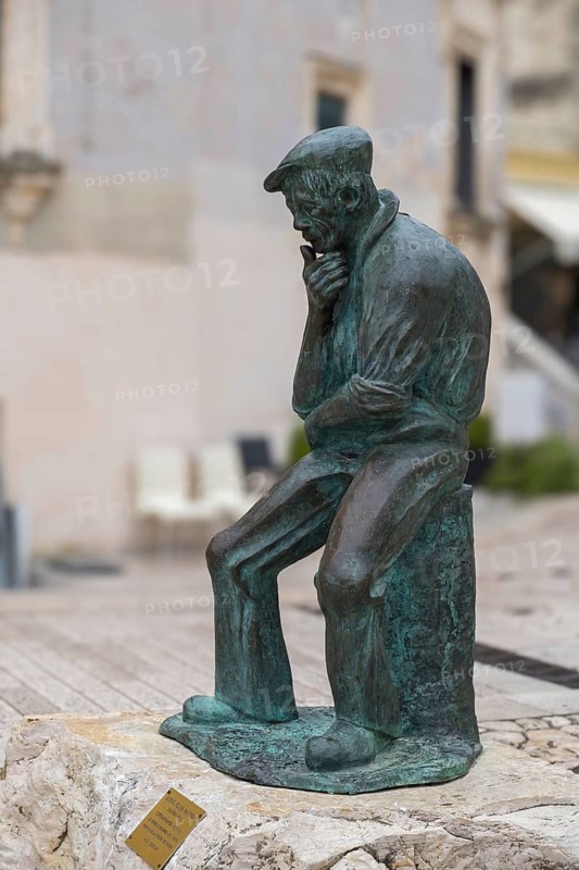 Bronze sculpture of a pensive man sitting on a stone in a historic city centre setting, Matera, Basalicata, Italy