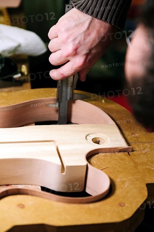 A luthier's hand carefully measures a bass guitar body with a caliper in a workshop, highlighting the detailed craftsmanship and precision in guitar making