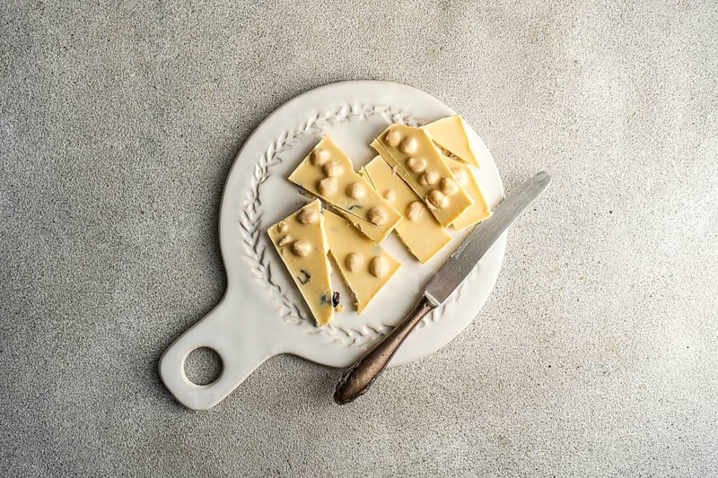 Top view of a white chocolate bar with hazelnuts, broken into pieces on a vintage white plate alongside a rustic knife on a textured grey concrete background