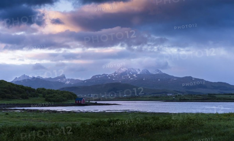 Landscape on the Lofoten Islands with meadows, sea and a small red hut, with other houses and mountains in the background. At night at the time of the midnight sun. Some coloured clouds in the sky. Lofoten, Norway