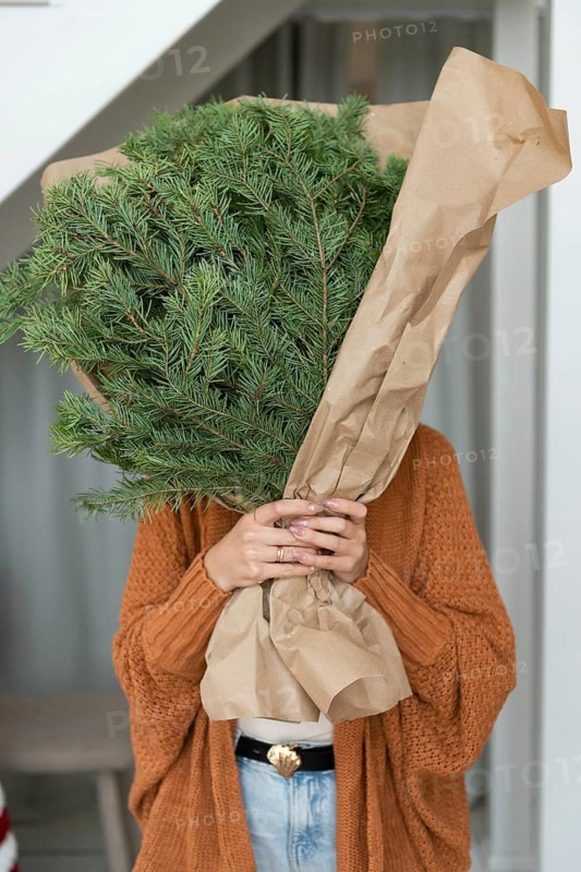 Unrecognizable female carrying a Christmas tree bouquet leaves covered in brown paper, representing holiday preparations