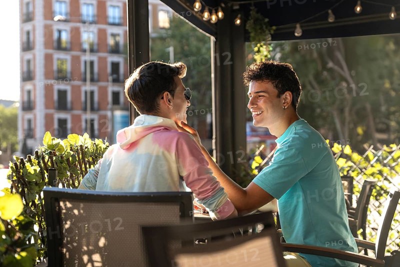 A joyful gay couple sitting at an outdoor cafe, sharing a tender moment. Sunlight filters through leaves, creating a warm, inviting atmosphere, highlighting their smiles