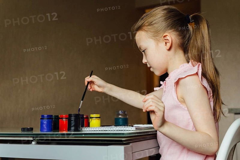 A young girl in a pink dress paints with colorful jars on a table. Engaged and focused, she explores her creativity in a cozy indoor setting
