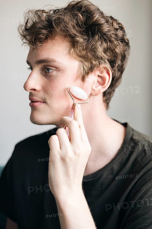 A young male relaxes at home while using a pink quartz facial roller on his skin, focusing on skincare and well-being