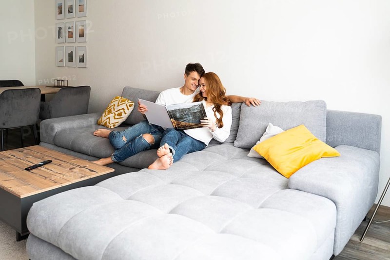 A joyful young couple relaxes on a cozy gray sofa, engaged in reading a large photo album. The warm ambiance highlights their excitement and anticipation for a new baby