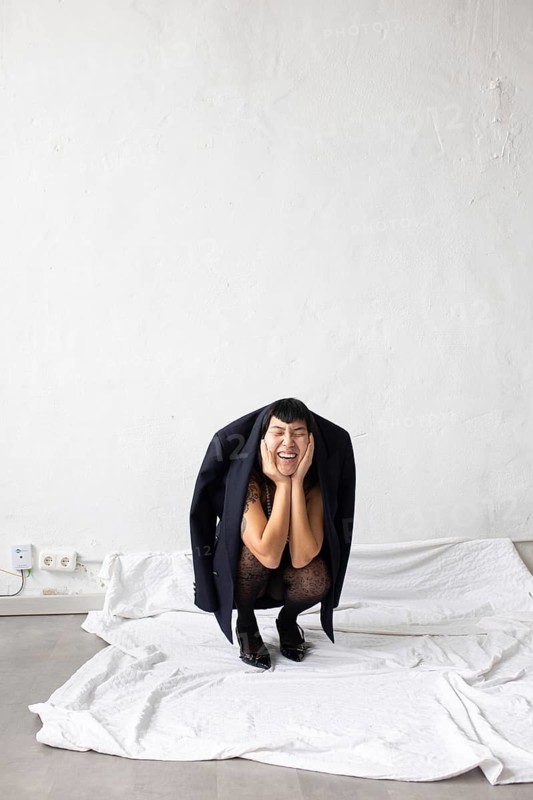 A joyful Asian woman is playfully crouching, wearing an oversized jacket. She expresses exuberance and charm in a minimalist setting, surrounded by white fabric