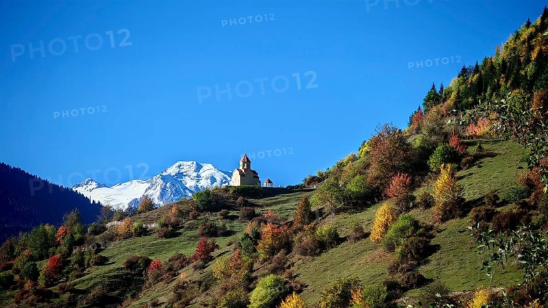 A church on a hill between colorful autumn trees, snow-capped peaks under a blue sky, Mestia, Svaneti, Georgia