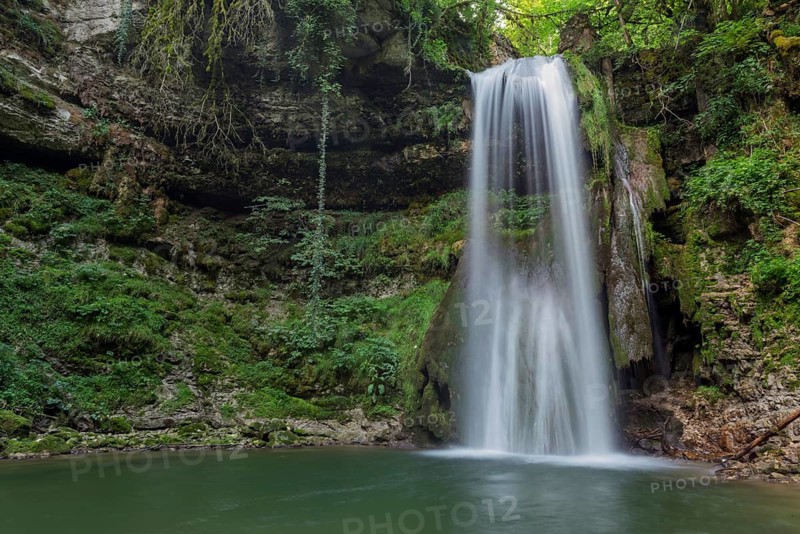 A waterfall flows into a small pool of water. The water is clear and the rocks are scattered around the pool. Lons le Saunier, Jura, Burgundy Franche Comte, France
