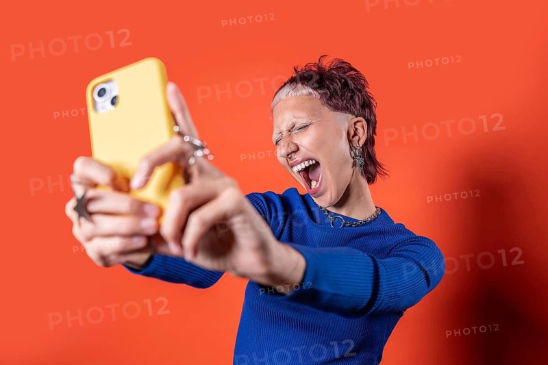 A young woman with a bold hairstyle, tooth gems and short bold hair takes a selfie with a yellow phone. Dressed in a blue sweater, she expresses excitement against a vibrant orange background