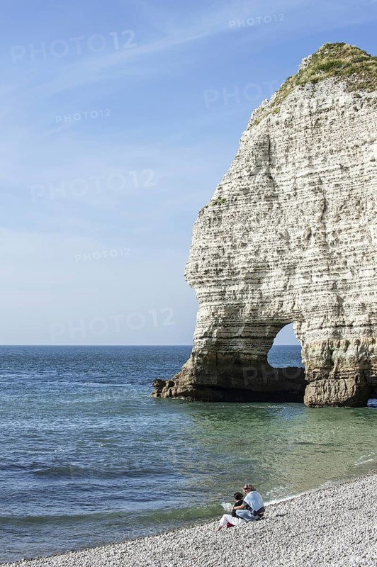 Couple sitting on pebble beach in front of the natural rock arch porte d'Amont at Etretat, Normandy, France