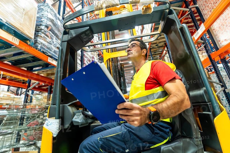 Side view of a concentrated mature man in forklift holding checklist in warehouse