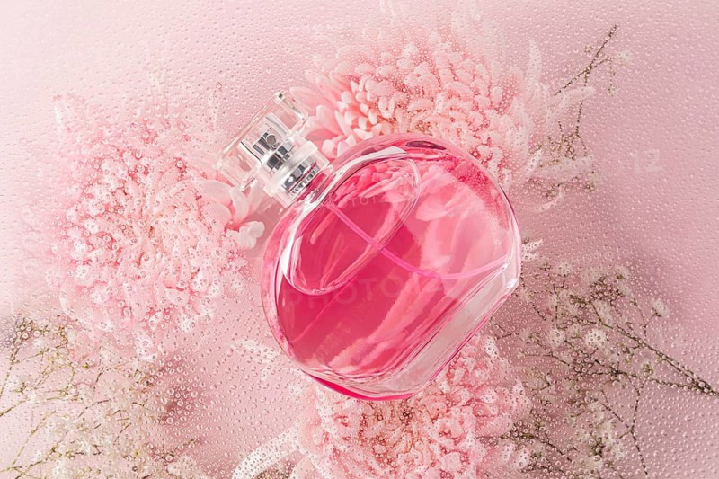 A sophisticated pink perfume bottle elegantly placed on a soft surface adorned with pink flowers and water droplets, exuding a sense of freshness and luxury