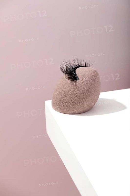 A false eyelash rests on a beige makeup sponge on a white surface, with a soft pastel background. The minimalist composition highlights the beauty and elegance of cosmetic tools