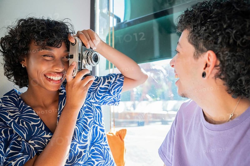A cheerful, multiethnic LGBTQ+ couple sharing a joyful moment as one partner playfully takes a photo of the other, capturing their happiness and togetherness indoors