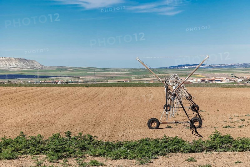 A center pivot irrigation system stands on freshly plowed farmland in Toledo, Spain. The rural landscape stretches toward distant hills and fields, showcasing agricultural infrastructure and open skies