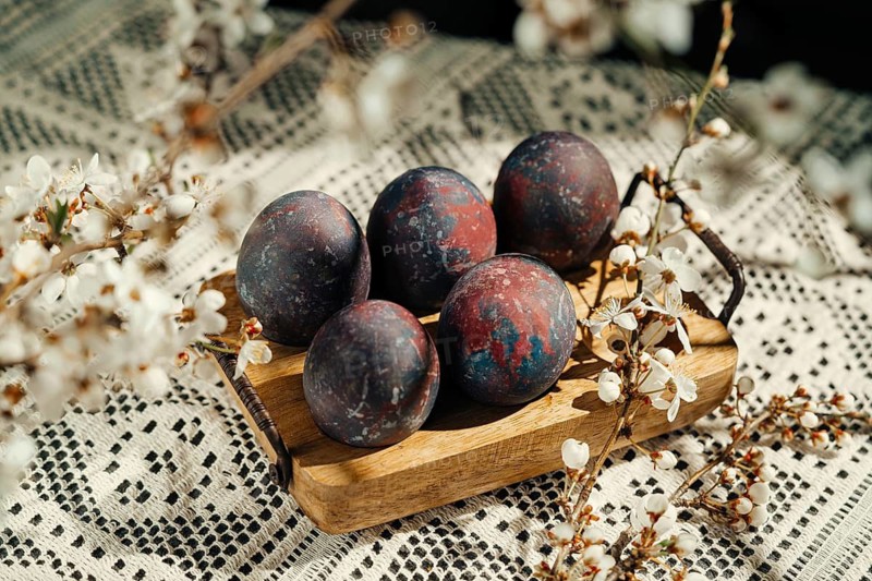 A rustic composition of four uniquely painted Easter eggs, nestled among delicate cherry blossoms on a wooden tray, lying over a textured crochet tablecloth. Perfect for spring and Easter themes