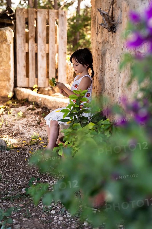 A young girl in a white dress sits by a rustic wall, surrounded by lush greenery and purple flowers. She holds sunflower leaves, deeply engrossed in nature, bathed in warm sunlight