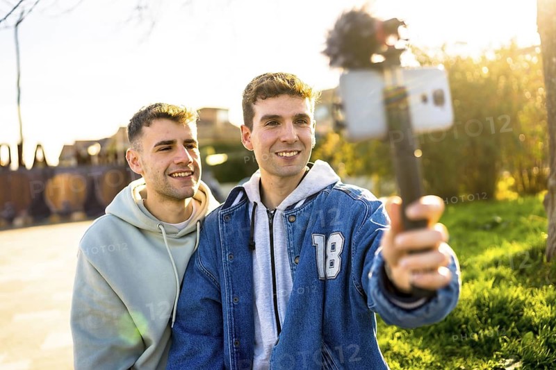 Two men filming a video blog outdoors, using a smartphone attached to a gimbal, smiling and enjoying their time together