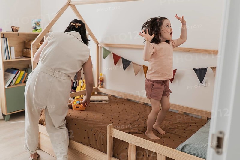A joyful moment of a mother and daughter together in a cozy bedroom. The mother tidies up while the daughter playfully jumps on the bed, surrounded by colorful toys
